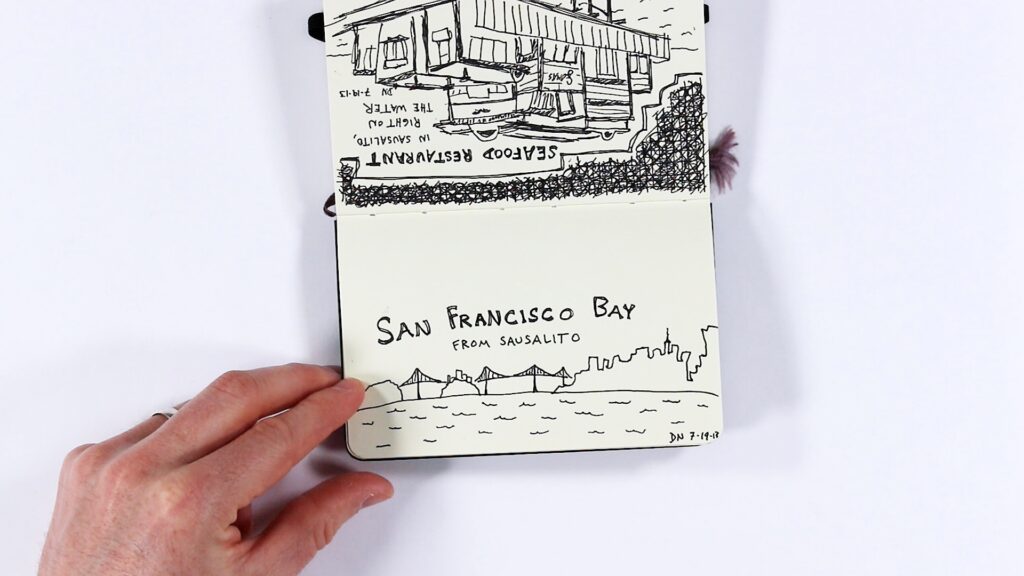 A hand holds open a small sketchbook showing two pen drawings. The top page (upside down) depicts a seafood restaurant in Sausalito, right on the water, with detailed line work of the building's structure. The bottom page shows a panoramic ink sketch of the San Francisco Bay as seen from Sausalito, featuring the city skyline, a bridge, and water, signed "DN 7-19-13."