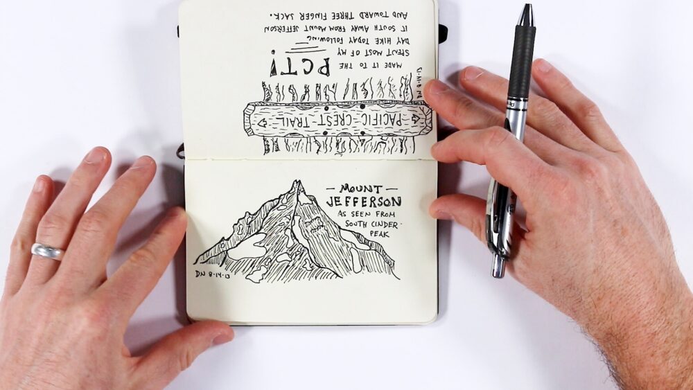 Two hands hold open a small sketchbook with a pen beside it. The top page (upside down) features a sketch of a Pacific Crest Trail sign post surrounded by trees, with text about spending most of the day hiking south away from Mount Jefferson and toward Three Finger Jack. The bottom page shows a detailed ink drawing of Mount Jefferson as seen from South Cinder Peak, with the mountain's jagged peaks rendered in fine line work, dated DN 8-14-13.