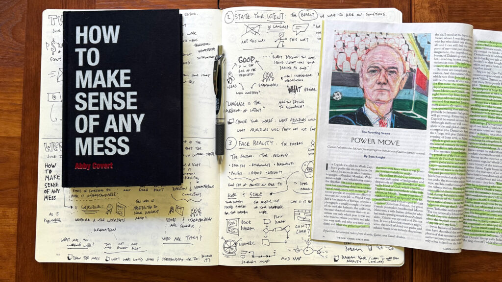 A flat lay photograph on a dark wooden table showing a reading and visual note-taking setup. At center-left is the black hardcover book "How to Make Sense of Any Mess" by Abby Covert. It rests on an open Moleskine-style sketchbook filled with dense handwritten sketchnotes — visual notes capturing key concepts from the book, including small icons, diagrams, hand-lettered headings like "State Your Intent," "Face Reality," and phrases about language, stakeholders, and information architecture. A black pen sits across the open pages. In the upper left corner, a small planner or notebook is partially visible with handwritten entries. To the right, a copy of The New Yorker magazine is open to an illustrated article titled "Power Move" by Sam Knight, featuring a painted portrait. The overall scene suggests a practice of slow, deliberate reading paired with visual sense-making — sketching out ideas by hand to better understand and remember them.